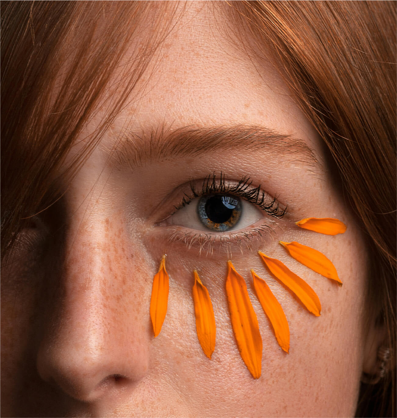 Zoom sur l’œil d’une femme aux cheveux roux avec des pétales orange délicatement placés sous le contour des yeux, symbolisant les bienfaits naturels des soins anti-cernes